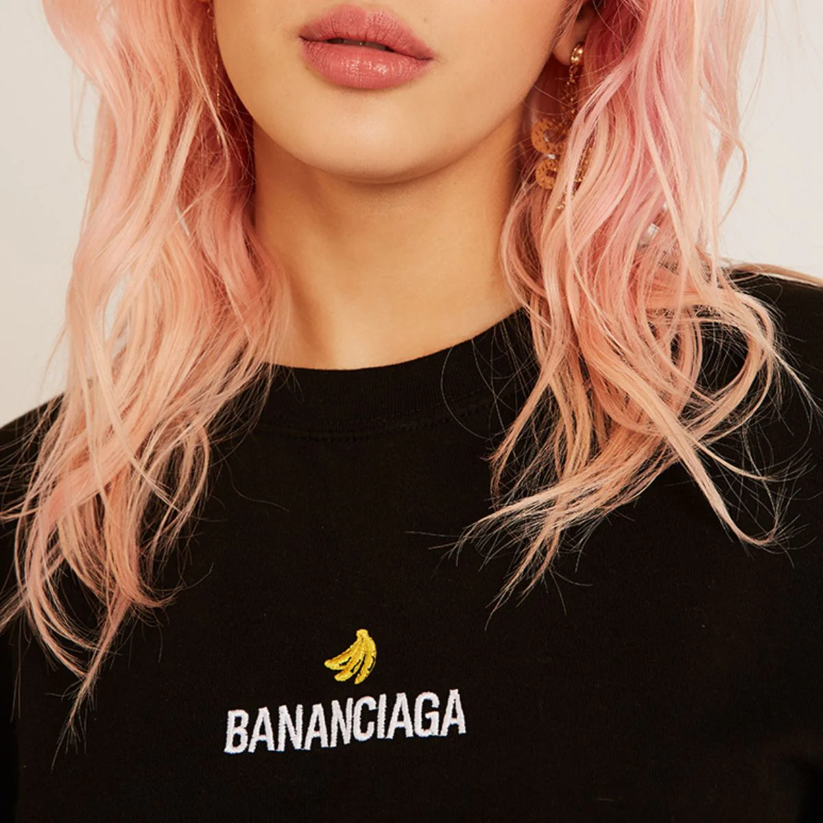 A person with wavy, pastel pink hair wears the Bananciaga Embroidered T-Shirt in black, featuring a yellow banana and "BANANCIAGA" in white—just the lower face and upper torso are shown, evoking chic Tallinn boutique style. Gate21.ee