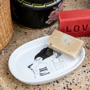 A bar of soap sits on the Bathroom - Tray, a white dish inspired by Estonian design and illustrated with two women, one brushing her hair. Paired with a red LOVE soap and a black floral container, it makes a charming display for gift or concept shops. Gate21.ee