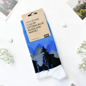 Eesti socks – blue-black-white forest: Blue, black, and white landscape socks with a brown "Why is the flag of Estonia blue black and white?" tag are shown on a white surface, surrounded by photos and small white flowers—a Tallinn shopping find. Gate21.ee
