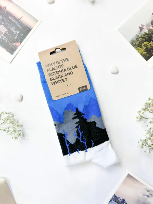 Eesti socks – blue-black-white forest: Blue, black, and white landscape socks with a brown "Why is the flag of Estonia blue black and white?" tag are shown on a white surface, surrounded by photos and small white flowers—a Tallinn shopping find. Gate21.ee