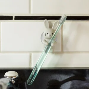 The Rabbit - Toothbrush Holder, a white ceramic bunny inspired by Estonian design, mounts on a tiled wall and holds a green toothbrush upright above a black sink worktop. Gate21.ee