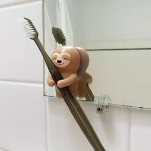 The Sloth - Toothbrush Holder, shaped like a sloth and made of plastic, clings to a bathroom mirror holding a brown toothbrush with white bristles. A second toothbrush stands in the background against white tiles. Gate21.ee