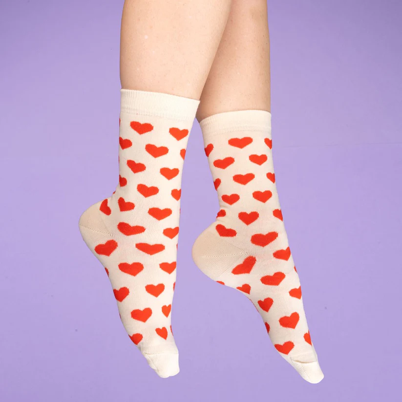 Feet wearing Hearts - Cotton Socks, featuring a red heart pattern on cream, positioned against a purple background—showcasing playful Estonian design. Gate21.ee