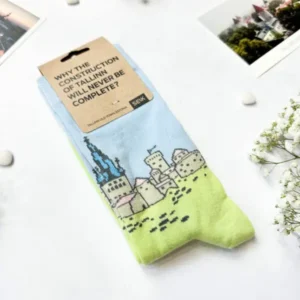 A pair of Eesti sokid – sini-must-valge mets socks, featuring a building and grass print, is curated with care on a white surface amid flowers and photos—a unique gift shop find. The tag reads: WHY THE CONSTRUCTION OF A LINE WILL NEVER BE COMPLETE?. Gate21.ee