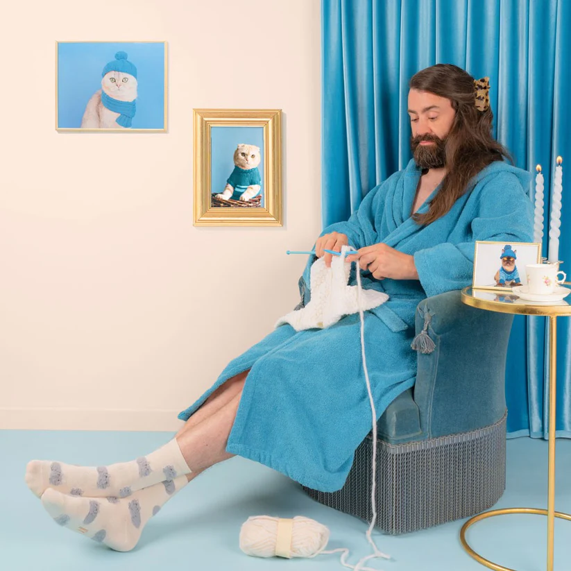 Wearing a blue dressing gown and Persian Cats - White Cotton Socks, a bearded person with long hair sits knitting in a blue armchair. The space resembles a curated gift shop, featuring cat portraits and creative touches like blue candles on the table. Gate21.ee