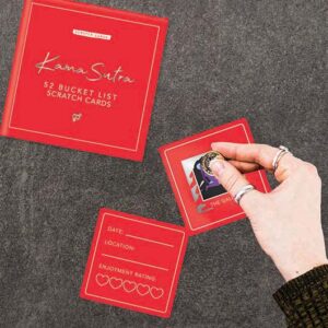 A hand with rings scratches a coin on a red card from the Kama Sutra - Scratch Cards Bucket List, with its red booklet and another card nearby&mdash;an Estonian-designed gift for sharing unique experiences. Gate21.ee