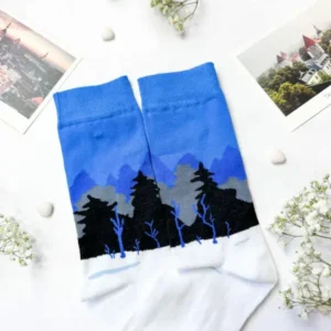 A pair of Eesti socks – sini-must-valge mets with a blue, black, and white forest design is displayed on a white surface next to a New Rebels small rucksack, surrounded by small white flowers, pebbles, and two landscape postcards. Gate21.ee