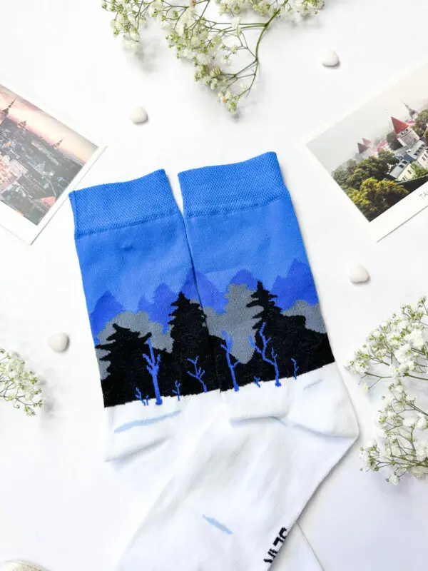 A pair of Eesti socks – sini-must-valge mets with a blue, black, and white forest design is displayed on a white surface next to a New Rebels small rucksack, surrounded by small white flowers, pebbles, and two landscape postcards. Gate21.ee