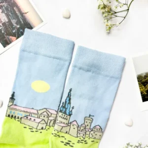 Eesti sokid – Tallinna vanalinn: Light blue socks with a yellow sun and townscape design, laid flat among flowers, pebbles, photos, and a New Rebels small rucksack—an ideal accessory combination. Gate21.ee