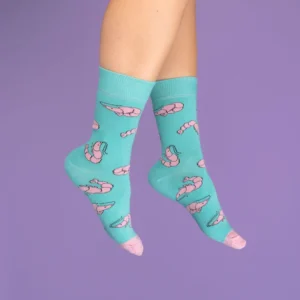 A pair of feet wear Shrimp - Cotton Socks, featuring a teal base with pink prawn designs and light pink toes and heels, set against a solid purple background. Gate21.ee