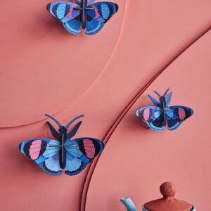 Three Peacock Butterflies from a set of 3 are displayed on a textured pink wall. A blue teapot with a red lid sits at the bottom right, creating a playful scene that’s ideal for those looking for unique Tallinn shopping ideas. Gate21.ee