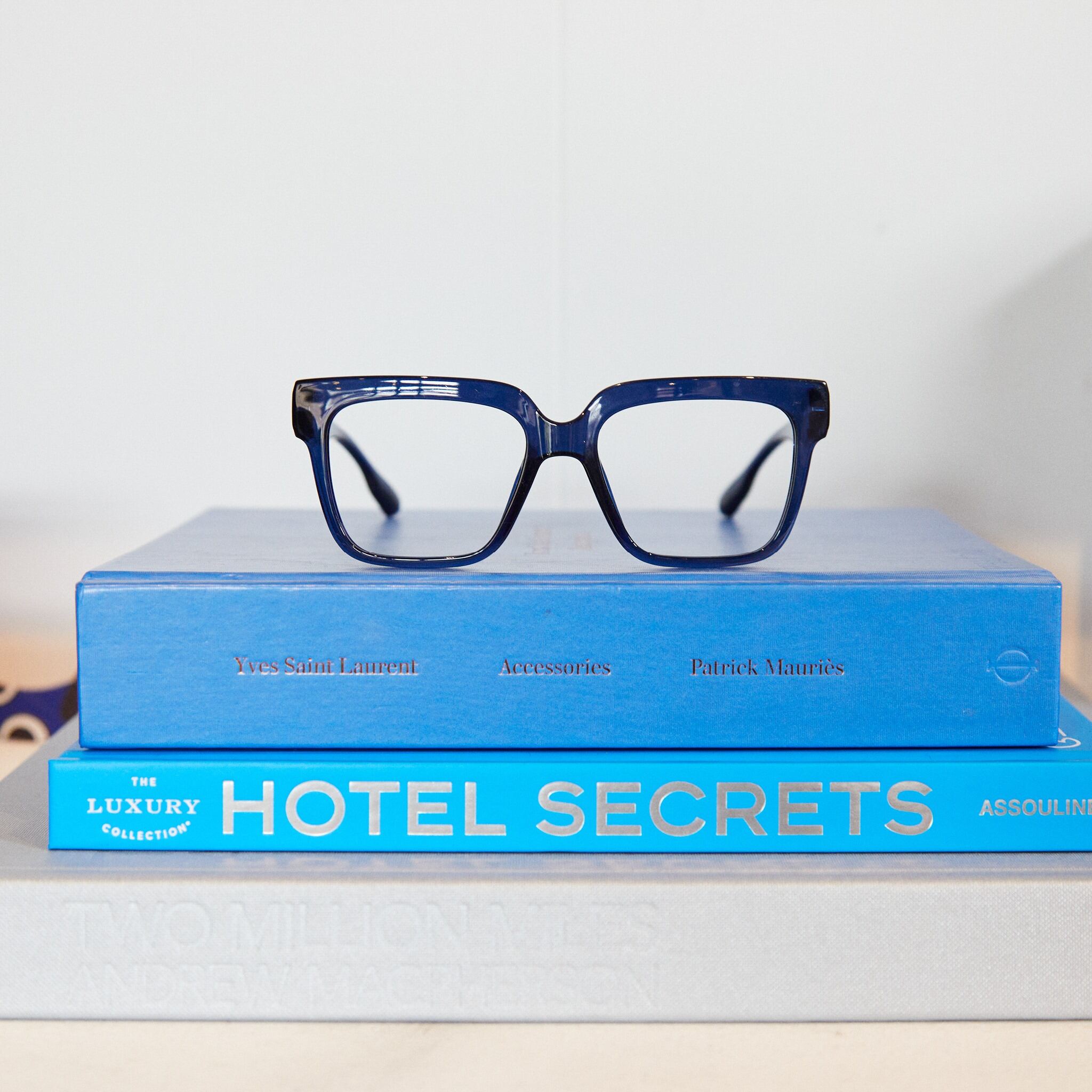 The MAX - Hazel dark blue eyeglasses rest atop two stacked blue books, "Yves Saint Laurent Accessories" and "Hotel Secrets," with a grey book beneath—an inspiring display for any concept shop. Gate21.ee