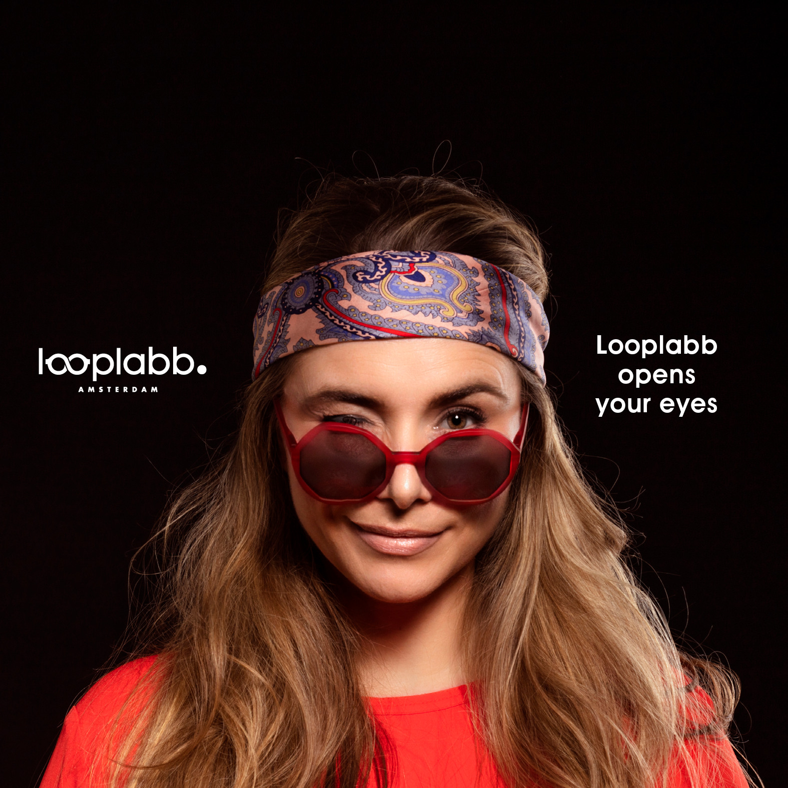 A woman with long hair wears THE GEORGE - Crystal Clear sunglasses and a paisley headband, winking at the camera. Dressed in red, she embodies Estonian design. The text reads: Looplabb Amsterdam and Looplabb opens your eyes. Gate21.ee