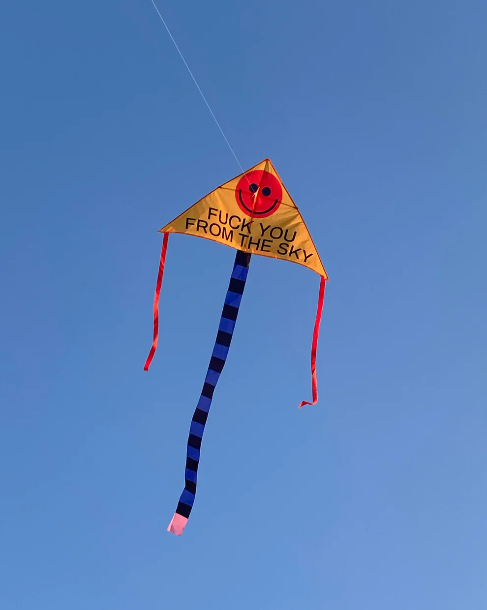 FUCK YOU FROM THE SKY - Kite: This bold yellow triangle kite features a red smiley face, playful message, striped tail, and red ribbons. Show off Estonian design with this quirky find for Tallinn shopping or any unique gift shop. Gate21.ee