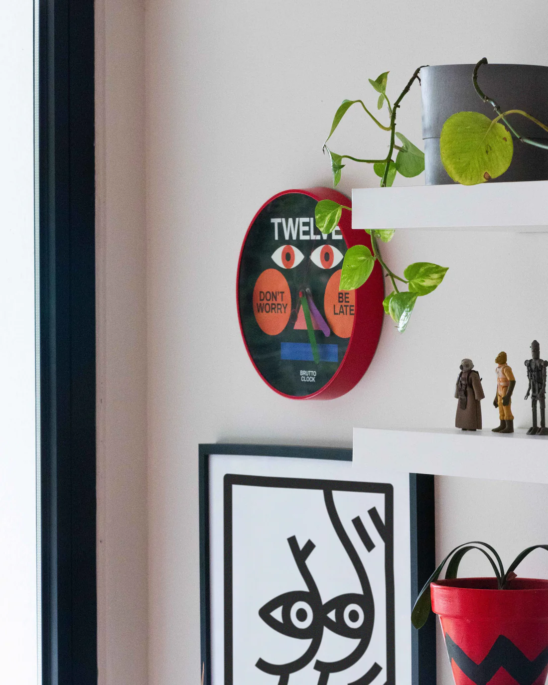 The BE LATE - Wall clock, featuring eyes and the text "DON'T WORRY" and "BE LATE," hangs above shelves with a potted plant, small figurines, and a framed abstract face—perfect for a concept shop full of unique ideas. Gate21.ee