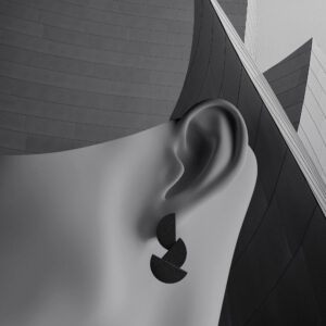 A greyscale image showing a close-up of a person's ear wearing a geometric, black, double-disc earring, set against a modern architectural background&mdash;perfect for a curated concept shop highlighting unique ideas in design. Gate21.ee