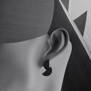 A greyscale image shows a close-up of a person’s ear wearing geometric, matte black earrings inspired by Estonian design, with a modern building featuring sleek, angular lines in the background. Gate21.ee