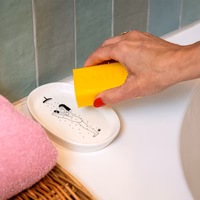 A hand with red nail varnish holds a yellow sponge, cleaning the 'Yoga in the Shower - Tray'—an ideal, quirky gift or souvenir to pick up while exploring local shops on your Tallinn holiday. Gate21.ee