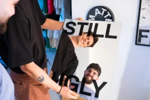 Two people hold a “STILL UGLY” mirror, their smiling reflections visible. A BE LATE wall clock hangs nearby, complementing curated decor and clothing that evoke the eclectic atmosphere of a unique gift shop. Gate21.ee