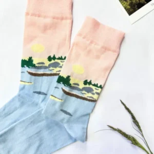 Eesti socks – Come to the island! pastel pink and blue landscape socks with sun, trees, and water details are laid flat on a white surface beside two photos, a sprig of grass, and a New Rebels small rucksack. Gate21.ee