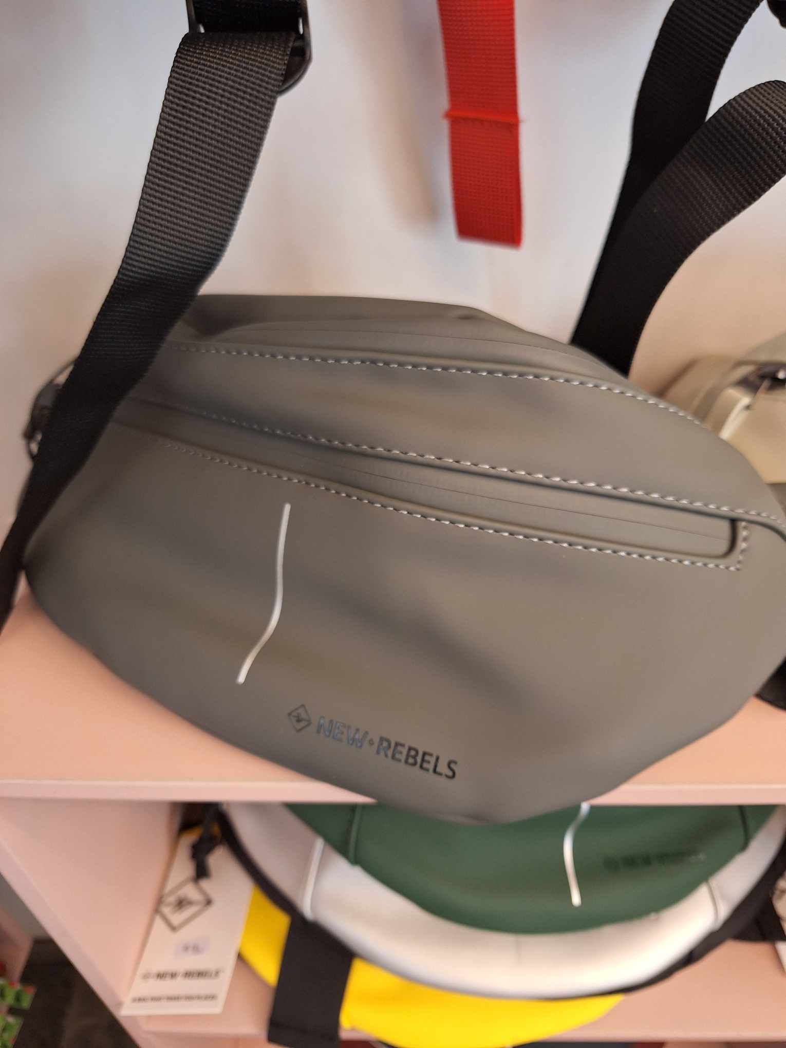 The Waistbag Vancouver - Anthracite by New Rebels, featuring a black strap and white zip detailing, is displayed on a shelf in a concept shop with other assorted bags visible in the background. Gate21.ee