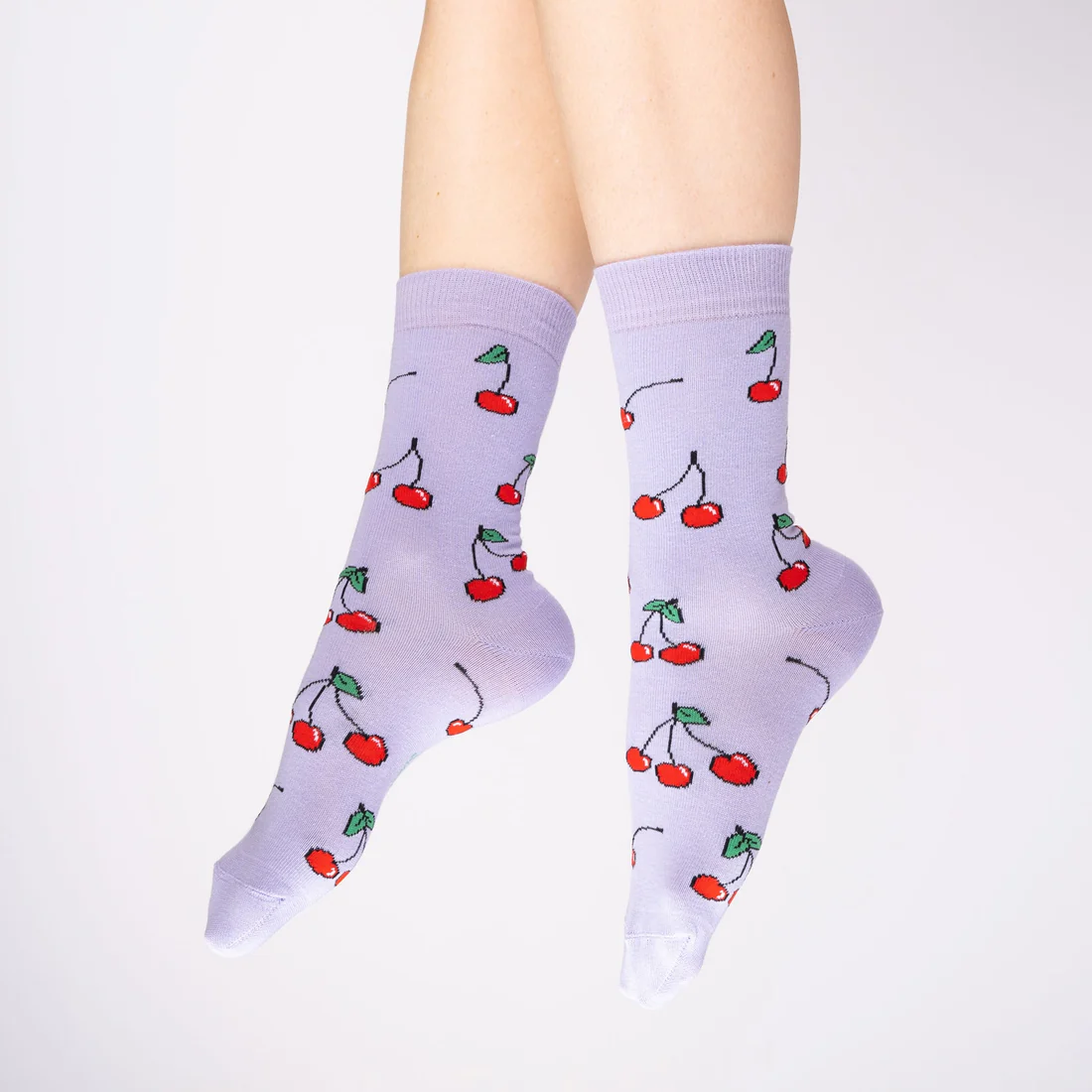 A pair of feet wears Cherries - Cotton Socks, featuring a red cherry pattern on light purple fabric, inspired by Estonian design, against a plain white background. Gate21.ee