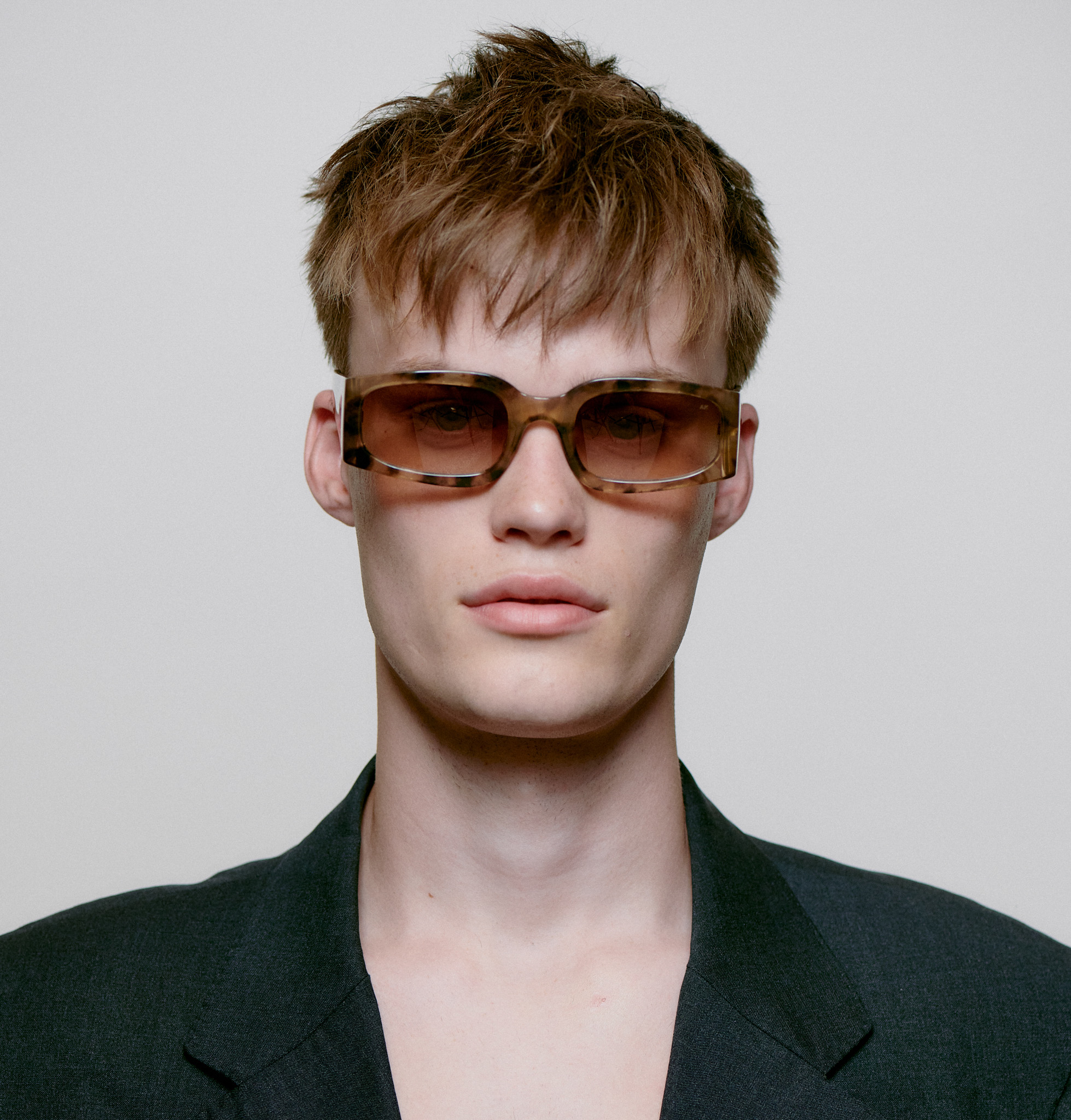 Wearing the ALEX - Coquina rectangular brown sunglasses, a person with short, tousled light brown hair and a dark jacket over a bare chest stands against a plain light background, channelling concept store style. Gate21.ee