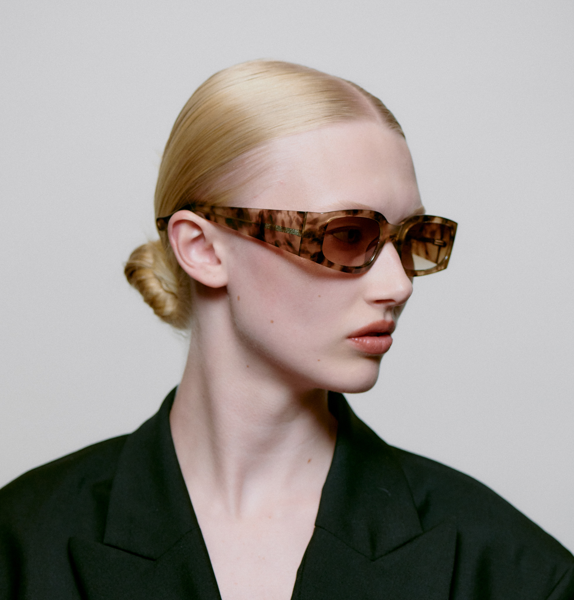 A person with light skin and blonde hair in a low bun wears ALEX - Coquina oversized brown tortoiseshell sunglasses and a black blazer, embodying chic Estonian design whilst looking to the side against a plain light background. Gate21.ee