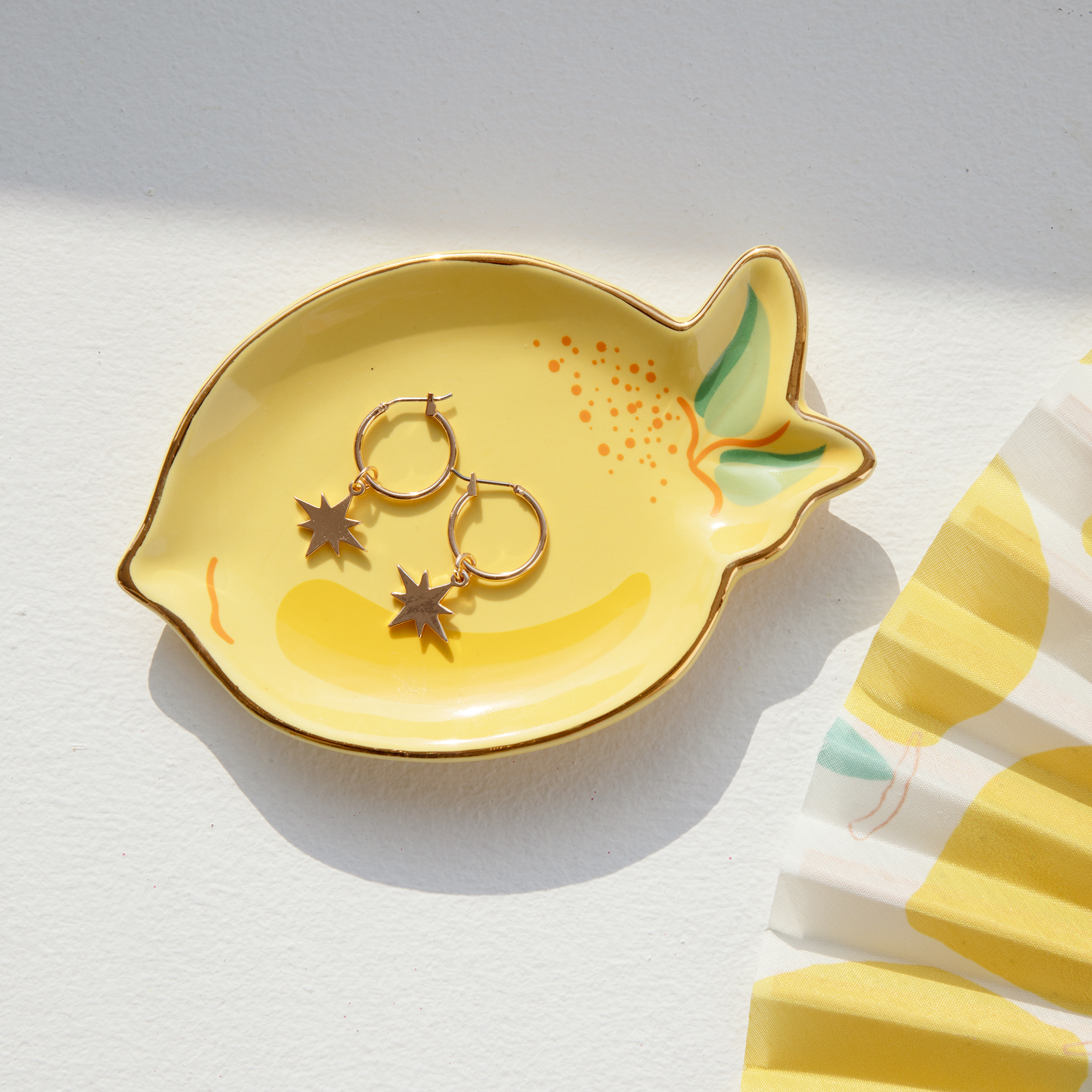 A pair of gold star-shaped hoop earrings sits on the Lemon - Jewellery Plate, a yellow ceramic dish with green accents. A yellow and white striped fabric peeks in, all arranged on a white surface. Gate21.ee