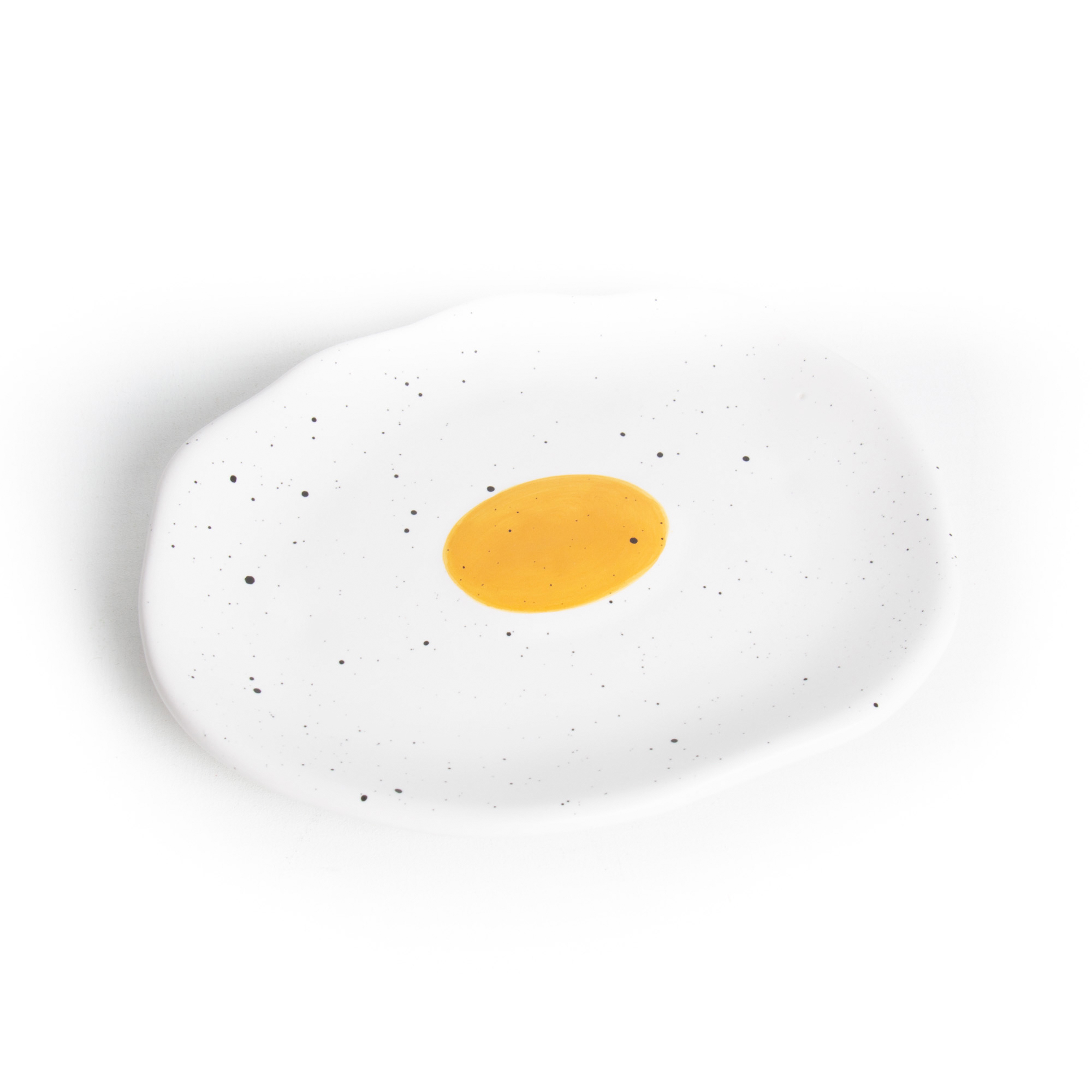 The Fried Egg - Jewellery Plate is a white ceramic plate with an irregular shape, black speckles, and a yellow circle in the centre—perfect as a unique Tallinn shopping find or to add charm to your gift shop. Background is white. Gate21.ee