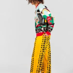 A person with curly hair stands sideways wearing the Trafico Jumper, featuring colourful Estonian-inspired graphic patterns, loose yellow trousers with black designs, and bright pink shoes against a plain white background. Gate21.ee