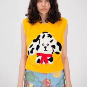 A woman with wavy brown hair wears the Manolito Waistcoat&mdash;a sleeveless yellow top with a cartoon dog, black spots, and red bow&mdash;paired with colourful trousers covered in playful illustrations, ideal for an Estonian design concept shop. Gate21.ee
