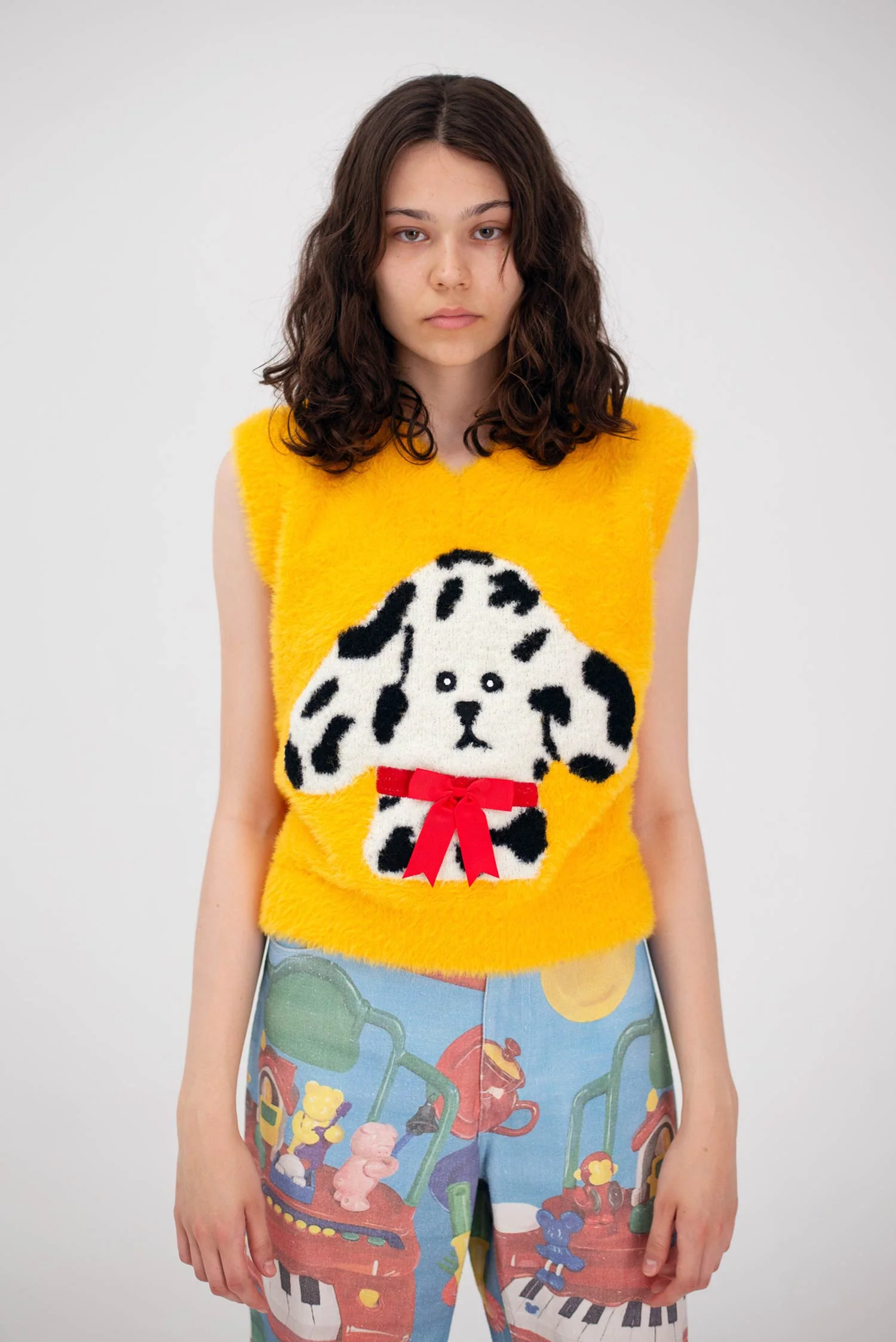 A woman with wavy brown hair wears the Manolito Waistcoat—a sleeveless yellow top with a cartoon dog, black spots, and red bow—paired with colourful trousers covered in playful illustrations, ideal for an Estonian design concept shop. Gate21.ee