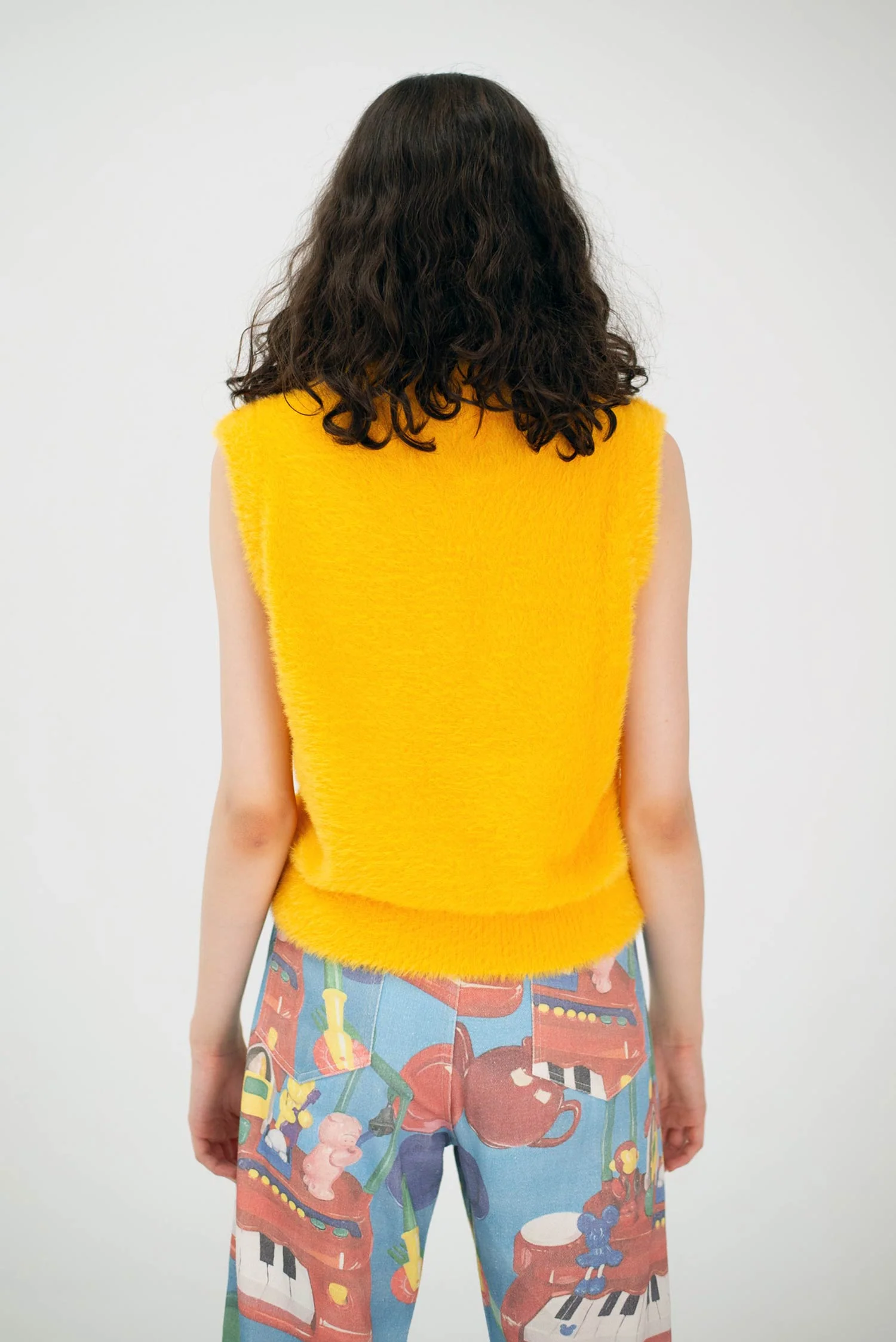 A person with shoulder-length curly brown hair is seen from behind, wearing the Manolito Vest—a sleeveless yellow fuzzy top—paired with colourful, whimsically printed trousers, for a unique look inspired by Estonian design and Tallinn shopping. Gate21.ee