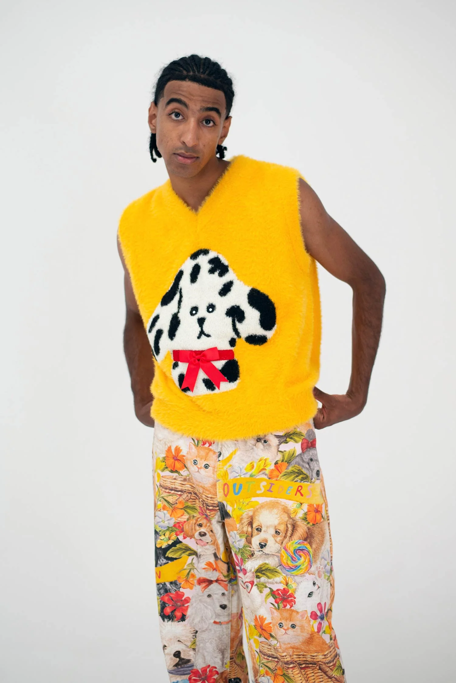 A person stands against a white background wearing the Manolito Vest—a bright yellow sleeveless top with a cartoon dog and red bow, showcasing playful Estonian design—paired with colourful shorts decorated with dogs, flowers, and "OUTSIDER. Gate21.ee