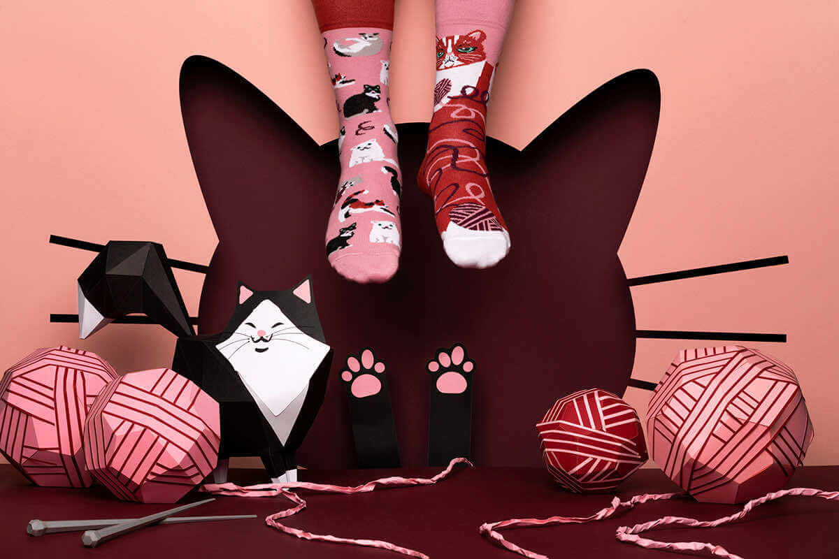 Two feet in cat-themed socks dangle above a dark cat-shaped cut-out. Pink and red balls of wool, knitting needles, and a paper cat sculpture from the Playful Cats collection highlight vibrant Estonian design against a pink backdrop. Gate21.ee