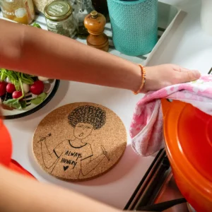 A person lifts the lid of an orange pot on the hob, their arm resting by a Helen B XL Coaster – Always Hungry Rita, adding a touch of Estonian design to this kitchen scene with fresh vegetables nearby. Gate21.ee