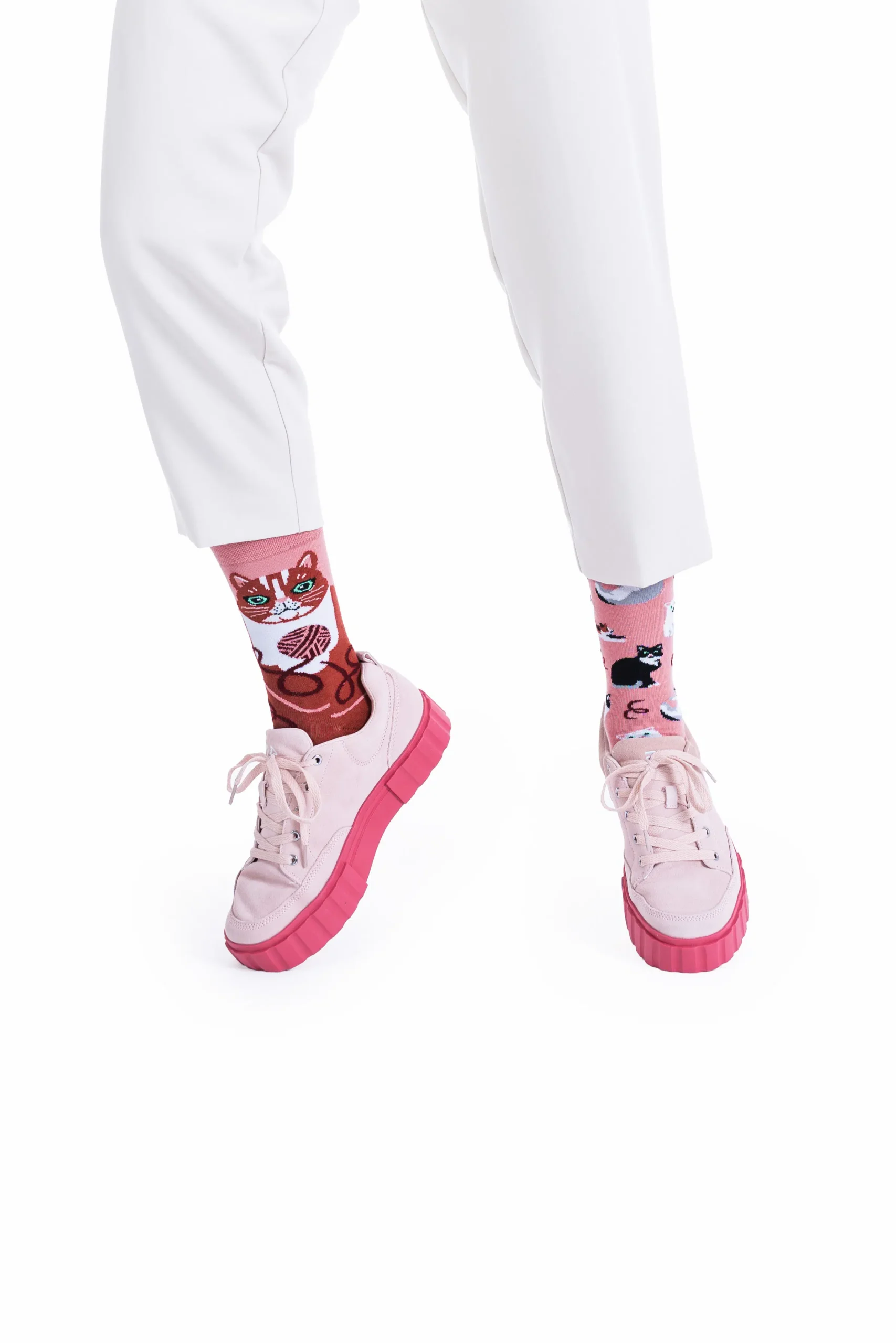 A person stands against a white background wearing white trousers and light pink trainers with thick pink soles, paired with Playful Cats mismatched pink socks featuring cat designs inspired by Estonian design. Gate21.ee