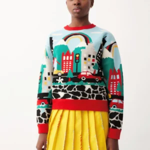 A person stands against a white backdrop wearing the Trafico Jumper, which features a colourful cityscape, rainbow, and car motif, paired with a yellow pleated skirt with silver accents. Gate21.ee