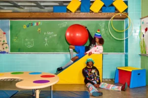 Two people in playful, colourful clothing pose in a classroom set with bright props. The curated scene highlights Estonian design and features the Spider Jumper, a yellow block, red ball, striped socks, and a whimsical chalkboard backdrop. Gate21.ee