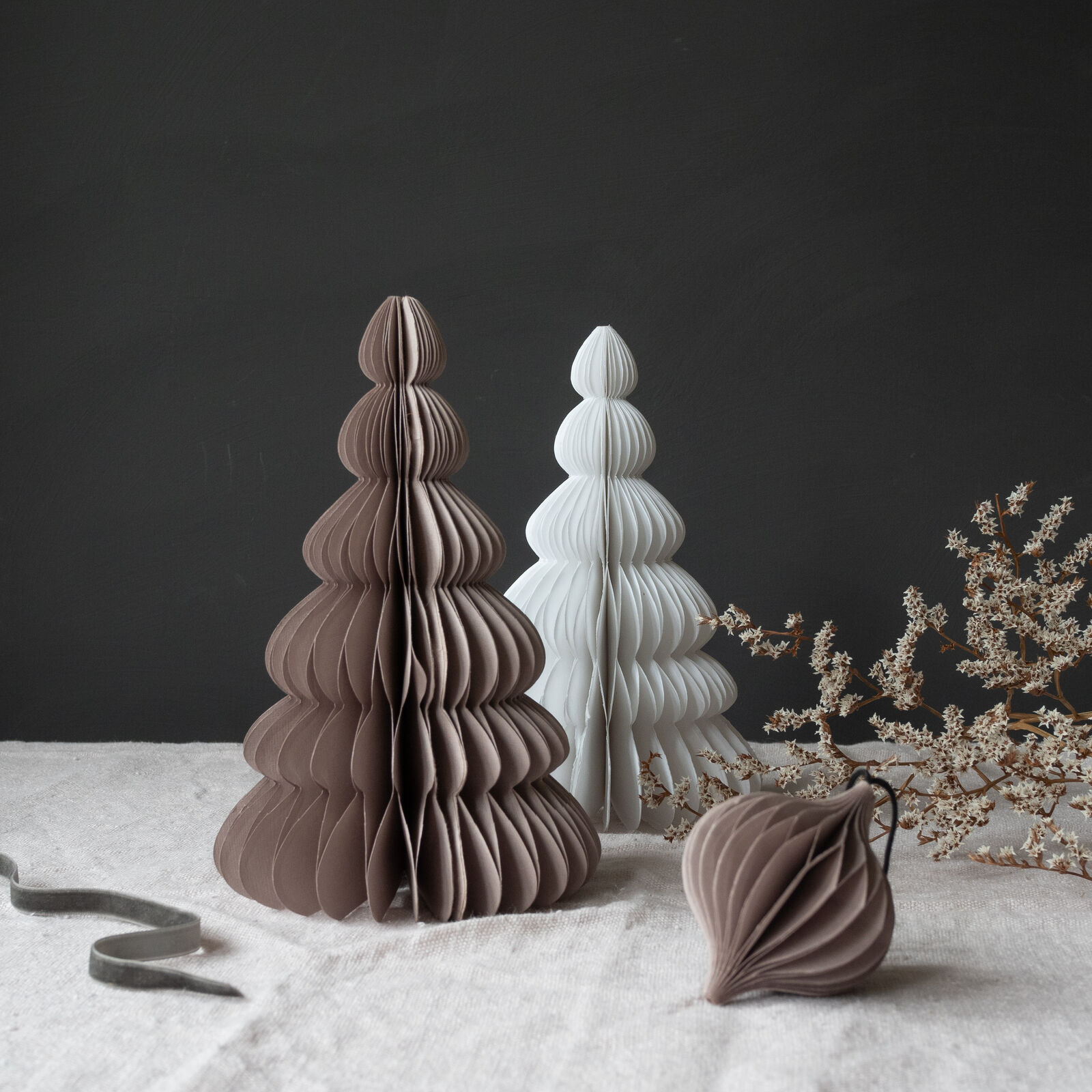 Two 30cm paper Christmas trees by KÕIK—one brown, one white—stand on light fabric beside a round bauble and dried branches. Perfect festive accents found at a Tallinn concept shop. Gate21.ee