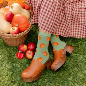 Wearing Squirrel - Cotton Socks with orange pumpkin designs, a person in a red gingham dress and brown clogs sits on grass beside a wicker basket filled with pumpkins and apples. Gate21.ee