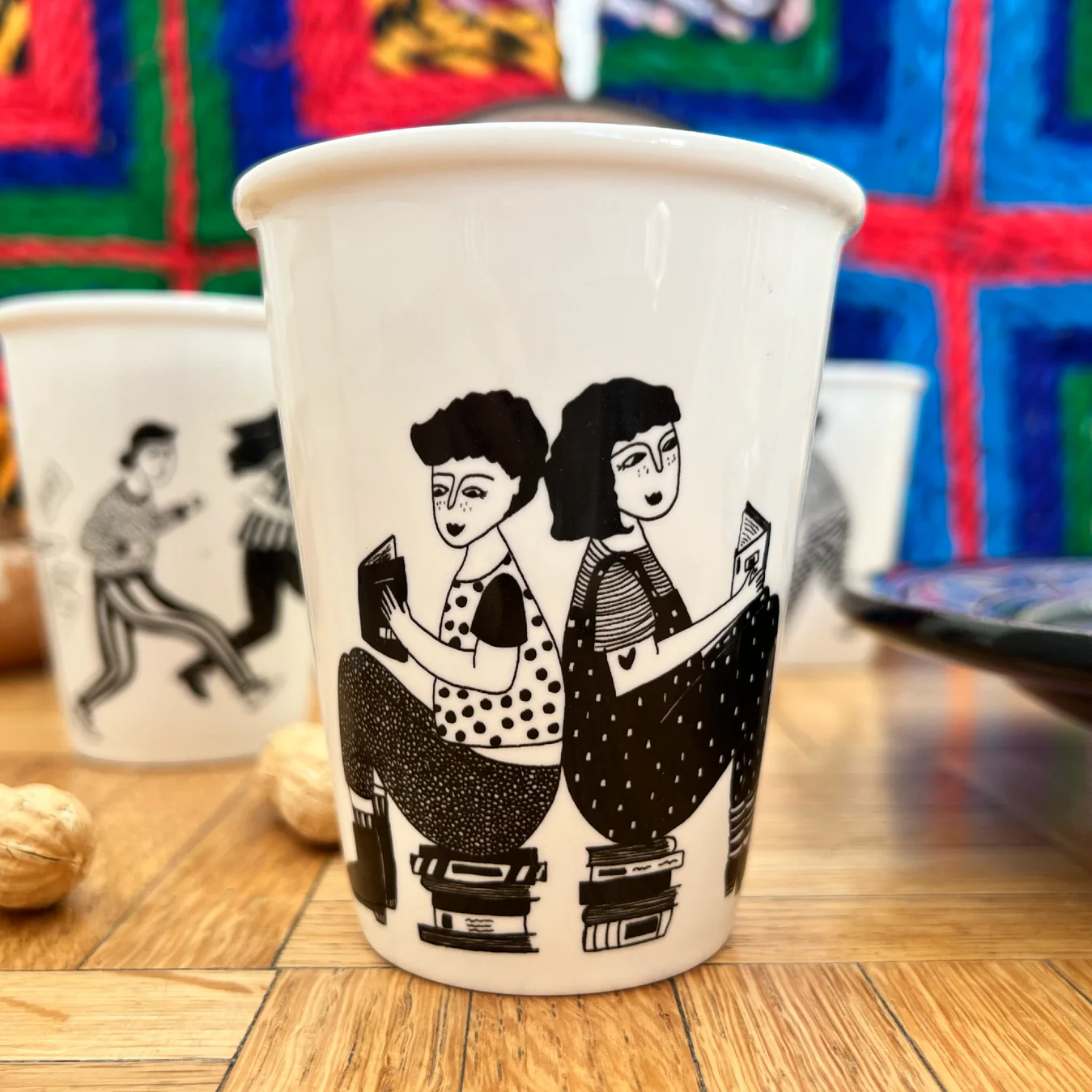 The Book Lovers Cup is a white mug with a black illustration of two people sitting back-to-back, each reading on stacks of books. Showcasing Estonian design, it’s pictured with other illustrated mugs, peanuts, and a colourful quilt in the background. Gate21.ee