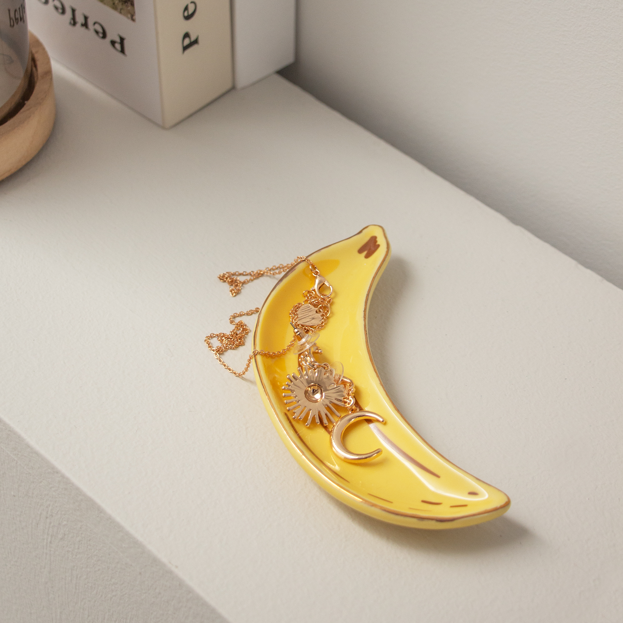 The Banana - Jewellery Plate, a yellow ceramic dish shaped like a banana, holds a gold sun and crescent moon necklace. Placed on a white surface beside books and a wooden coaster, it adds unique charm to your decor. Gate21.ee