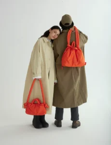 Two people in long coats stand against a plain background. One carries the Ona Soft Bag - Black by hand, whilst the other wears it over their shoulder, showcasing the curated simplicity and unique spirit of Estonian design. Gate21.ee