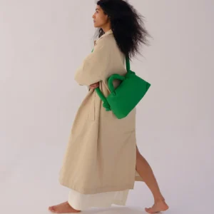 A person with long curly hair walks barefoot in a beige coat, carrying the Mini Ona Soft Bag - Green—perfect inspiration for unique finds on a Tallinn shopping trip or at a modern concept shop. Gate21.ee