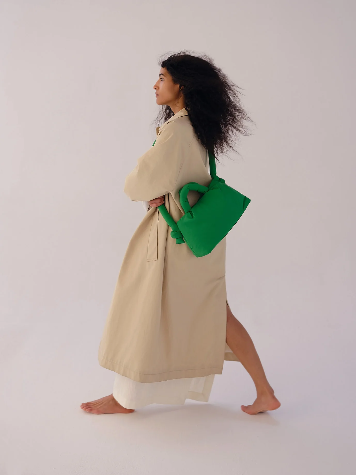 A person with long curly hair walks barefoot in a beige coat, carrying the Mini Ona Soft Bag - Green—perfect inspiration for unique finds on a Tallinn shopping trip or at a modern concept shop. Gate21.ee