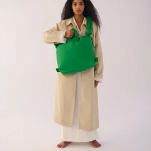 A person with long, curly hair stands barefoot against a plain background, wearing a beige coat over a white dress and holding the Ona Soft Bag - Green—a striking example of curated Estonian design. Gate21.ee