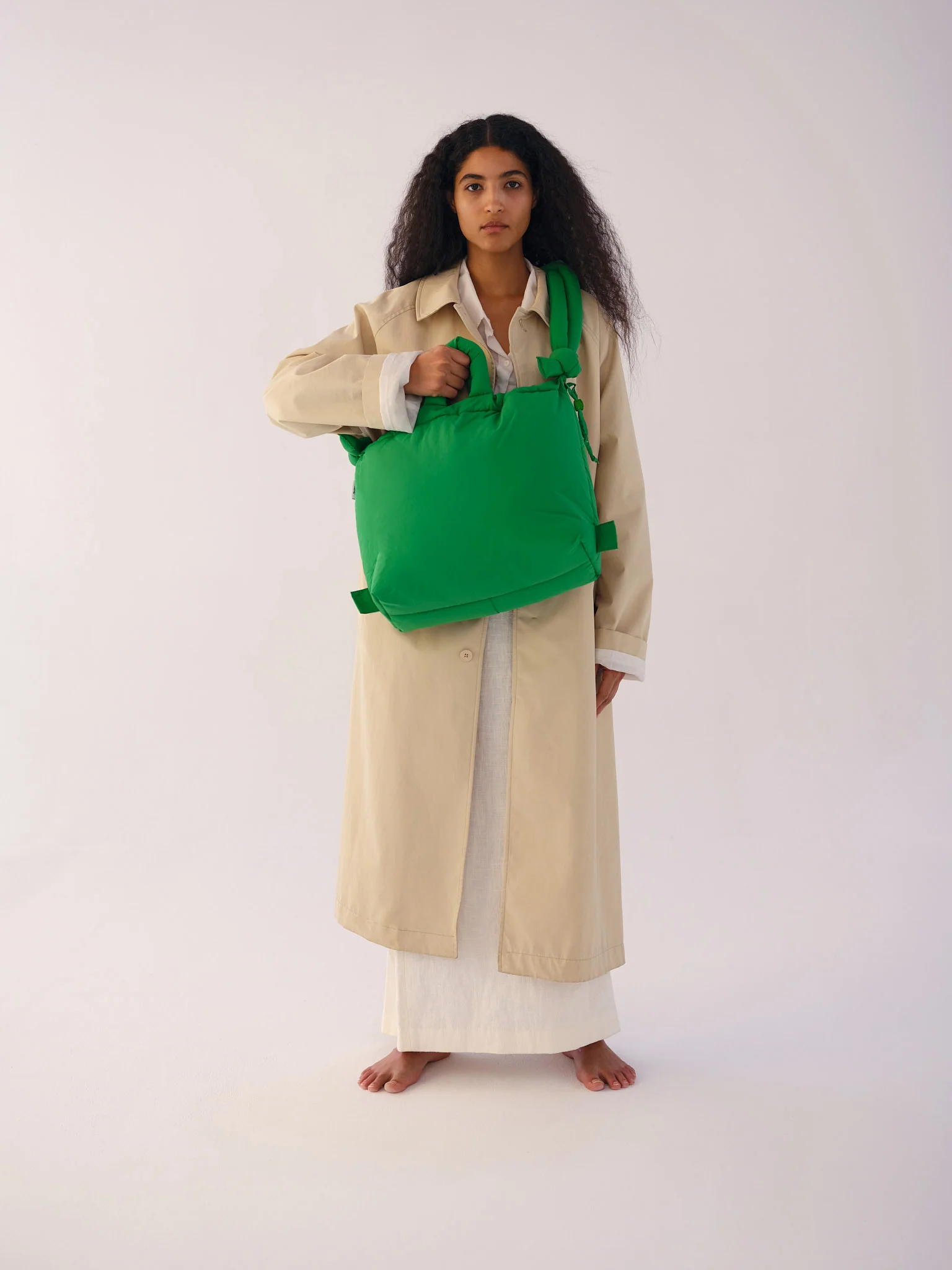 A person with long, curly hair stands barefoot against a plain background, wearing a beige coat over a white dress and holding the Ona Soft Bag - Green—a striking example of curated Estonian design. Gate21.ee