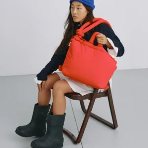 A person sits on a wooden chair, holding the Ona Soft Bag - Red—a large, bright red padded bag in stylish Estonian design. They’re dressed in a blue knitted hat, navy jumper, white dress, and black boots against a plain background. Gate21.ee